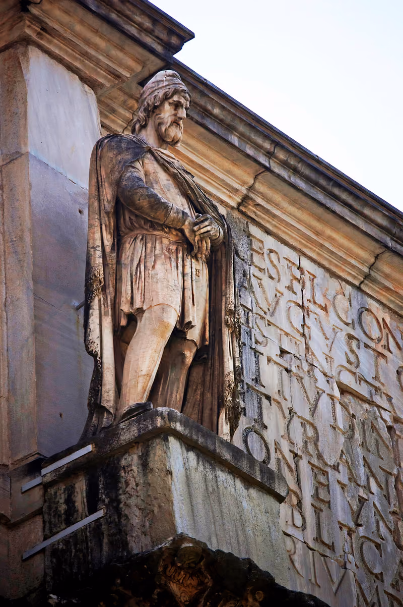 Statuie de marmură a unui dac, sus pe Arcul lui Constantin din Roma: bărbat cu barbă și plete, cu o căciulă pe cap, îmbrăcat cu tunică și mantie, cu mâinile împreunate în față.