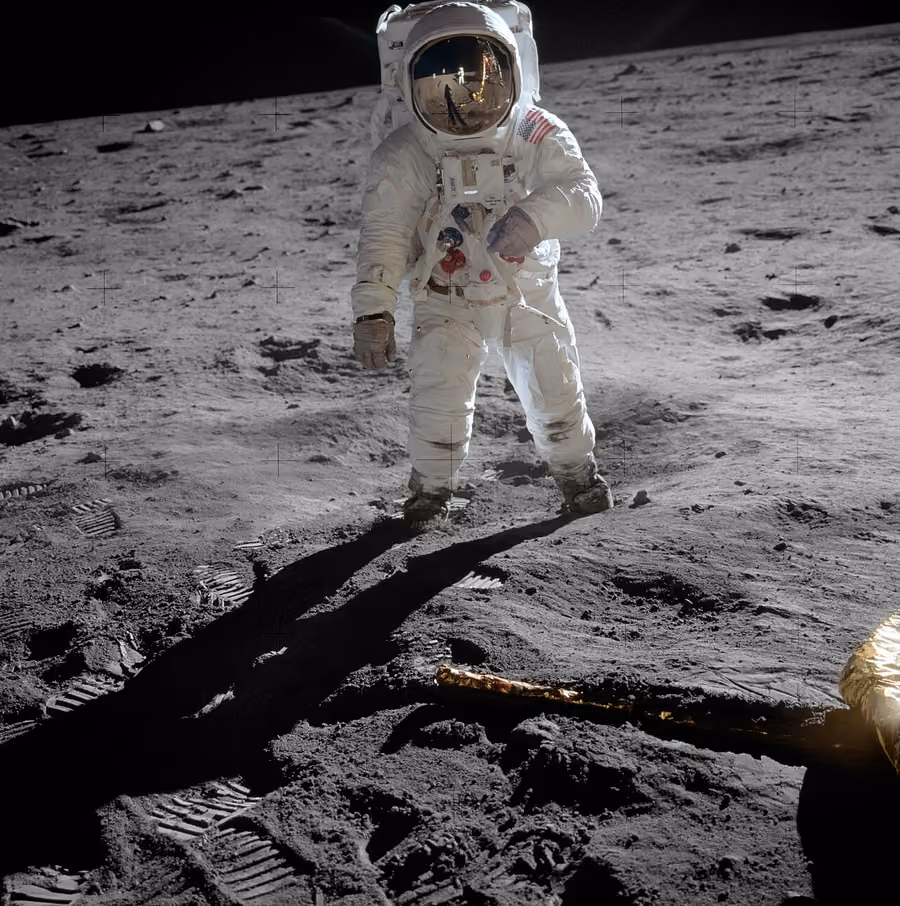 Astronaut în costum alb pe suprafața cenușie a Lunii, cu steagul SUA pe umăr; în vizieră se reflectă modulul lunar și un alt astronaut; se văd urme de pași în praf.