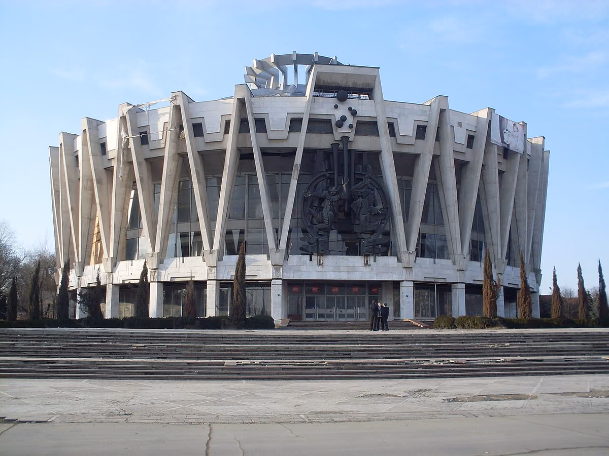 Clădire rotundă de beton, cenușie, cu stâlpi înclinați în formă de V în jurul fațadei, cu un relief sculptural întunecat în centru și trepte late în față.